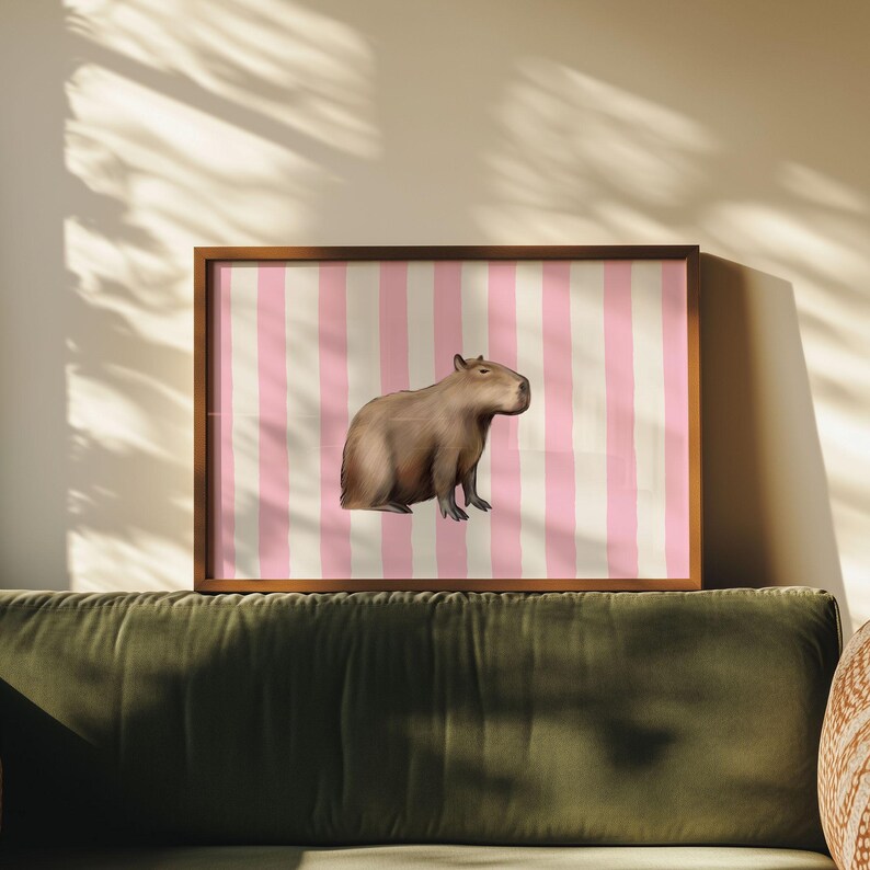 Op de afbeelding: Een ingelijste print van een capybara op een roze en witte gestreepte achtergrond. De capybara is bruin en kijkt naar rechts.
