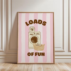 Op de afbeelding: Een ingelijste print met een roze en witte gestreepte achtergrond. Het kunstwerk toont een bruine illustratie van een wasmachine, een wasmand en de woorden "LOADS OF FUN" in bruine letters. De print heeft een houten lijst.