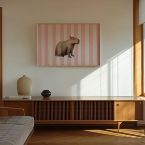 Op de afbeelding: Een bruin capibara zit op een roze en wit gestreepte achtergrond in een ingelijst schilderij dat aan een witte muur hangt. Op de voorgrond staat een houten kast met een lattenfront en een gesloten deur.