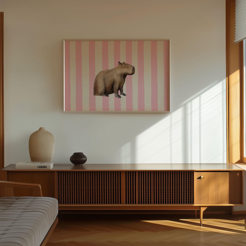 Op de afbeelding: Een bruin capibara zit op een roze en wit gestreepte achtergrond in een ingelijst schilderij dat aan een witte muur hangt. Op de voorgrond staat een houten kast met een lattenfront en een gesloten deur.