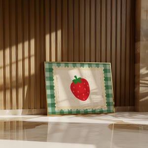 Puede incluir: Una impresi&oacute;n enmarcada con una fresa roja con hojas verdes. La obra de arte tiene un borde a cuadros verde y blanco y un marco dorado. El fondo es de color beige claro.