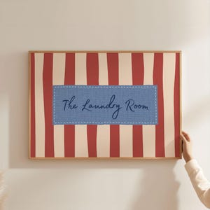 May include: A framed print with a red and cream striped pattern. The centre features a blue denim-style rectangle with the cursive text "The Laundry Room". The frame is a light brown colour.