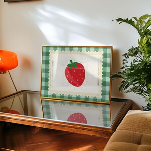 Puede incluir: Una impresi&oacute;n enmarcada con una ilustraci&oacute;n de fresa roja con tallo y semillas verdes. La obra de arte est&aacute; sobre un fondo blanco con un borde similar a un sello postal, enmarcada por un patr&oacute;n de cuadros verdes y blancos. El marco es dorado.