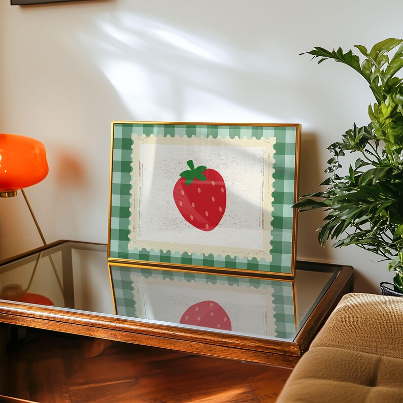 Puede incluir: Una impresi&oacute;n enmarcada con una ilustraci&oacute;n de fresa roja con tallo y semillas verdes. La obra de arte est&aacute; sobre un fondo blanco con un borde similar a un sello postal, enmarcada por un patr&oacute;n de cuadros verdes y blancos. El marco es dorado.
