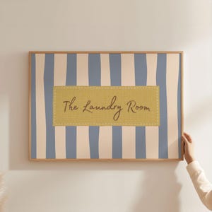 Puede incluir: Una obra de arte enmarcada con un marco de madera clara. La obra presenta rayas verticales en azul claro y blanco roto. Un parche rectangular con el texto "The Laundry Room" está centrado en la obra.