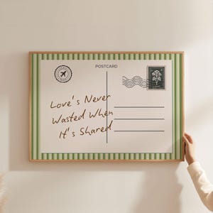 Puede incluir: Un diseño de postal enmarcado con un marco de madera. La postal presenta bordes a rayas verdes y blancas, un sello y el texto "Love's Never Wasted When It's Shared". Un logotipo con temática de viajes está en la esquina superior izquierda.