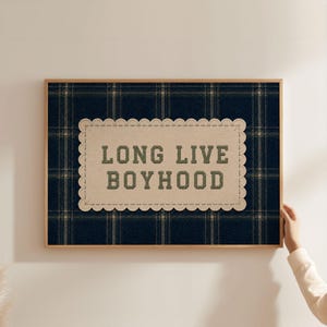 Könnte beinhalten: Ein rechteckiges Wandbild mit Rahmen und einem dunkelblauen sowie beigefarbenen Karomuster. In der Mitte befindet sich ein beiger Aufnäher mit gewelltem Rand und dem dunkelgrün gestickten Text "LONG LIVE BOYHOOD" in Blockschrift.