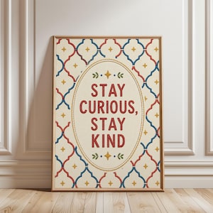 Könnte beinhalten: Gerahmter Druck mit den Worten "STAY CURIOUS, STAY KIND" in Rot, innerhalb eines ovalen Rahmens. Der Hintergrund zeigt ein cremefarbenes Muster mit roten, blauen und gelben geometrischen Formen. Der Rahmen ist hellbraun.
