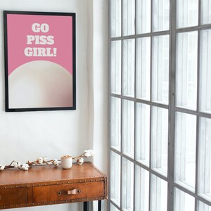 May include: A framed poster with a pink background and white text that reads "GO PISS GIRL!" The poster is hanging on a white wall next to a window with white blinds. A small wooden table with two drawers is in the foreground.