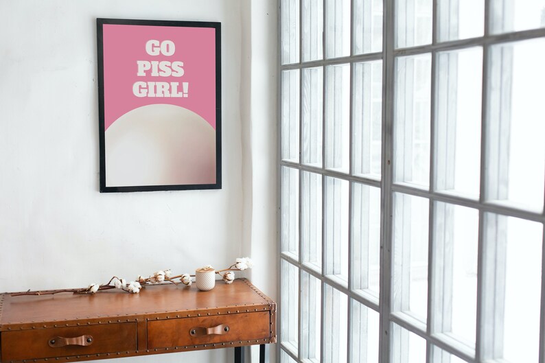 May include: A framed poster with a pink background and white text that reads "GO PISS GIRL!" The poster is hanging on a white wall next to a window with white blinds. A small wooden table with two drawers is in the foreground.