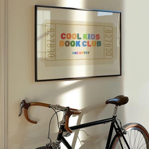 May include: A framed print with the text "COOL KIDS BOOK CLUB" in colorful letters. The print resembles a vintage ticket and is displayed above a black bicycle with brown handlebars and a brown leather seat.