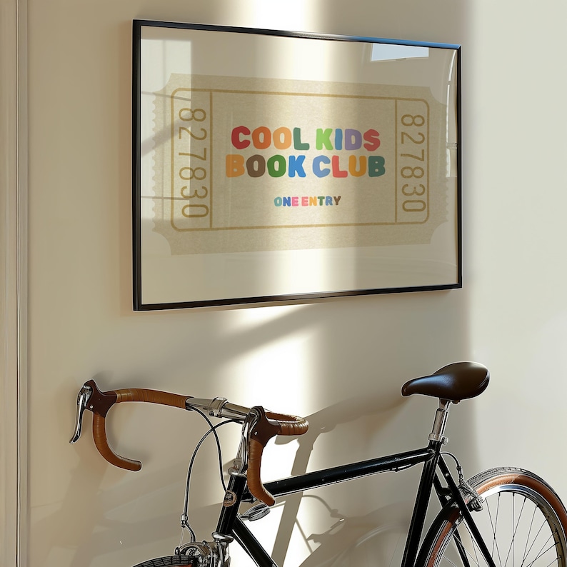 May include: A framed print with the text "COOL KIDS BOOK CLUB" in colorful letters. The print resembles a vintage ticket and is displayed above a black bicycle with brown handlebars and a brown leather seat.