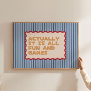 May include: A framed print featuring a blue striped background and the text "ACTUALLY IT IS ALL FUN AND GAMES" within a cream-coloured, scalloped-edged frame. The frame is a light brown colour.