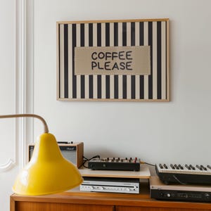 Puede incluir: Una obra de arte enmarcada con rayas verticales en blanco y negro y el texto "COFFEE PLEASE" en un parche beige. Una l&aacute;mpara amarilla, equipo de audio y un teclado se encuentran sobre un mueble de madera.