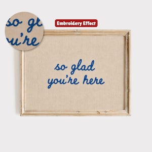 May include: A framed artwork with the phrase "so glad you're here" embroidered in blue thread on a beige background. The frame is a light brown color, and the artwork is displayed against a white background.