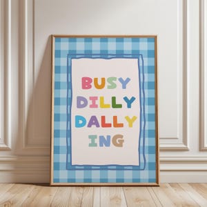 May include: A framed print with the words "BUSY DILLY DALLY ING" in colorful letters. The text is on a white background, framed by a blue gingham pattern and a thin blue border. The frame is a light brown color.