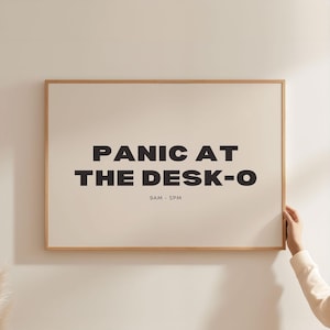 Könnte beinhalten: Ein gerahmtes Poster mit dem Text "PANIC AT THE DESK-O" in fetten schwarzen Buchstaben. Unter dem Text steht "9AM - 5PM" in kleinerer Schrift. Der Rahmen ist hellbraun und der Hintergrund cremefarben.