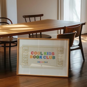 May include: A framed print featuring a ticket design with the text "COOL KIDS BOOK CLUB" in colorful letters. The print has a light beige background and is displayed on a wooden floor, with a wooden table and chairs in the background.