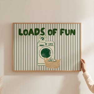 May include: A framed print with the words "LOADS OF FUN" in green block letters. The artwork features a washing machine illustration with a basket of laundry. The background has green and white vertical stripes.