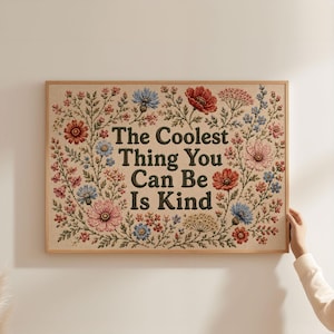 May include: A rectangular wall art piece in a light wood frame featuring a beige background with a floral border of red, pink, and blue flowers. The center text in dark brown, bold, serif font reads: The Coolest Thing You Can Be Is Kind.