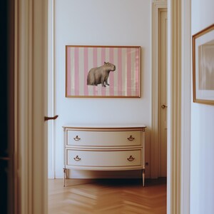 Op de afbeelding: Een witte commode met twee laden en gouden accenten staat in een gang met een ingelijste print van een capibara op roze en witte strepen op de muur.
