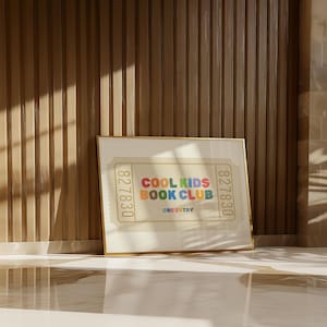 May include: A framed print with the text "COOL KIDS BOOK CLUB" in colorful letters. The print has a ticket design with the number "827830" on each side. The frame is gold, and the background is a light beige color.
