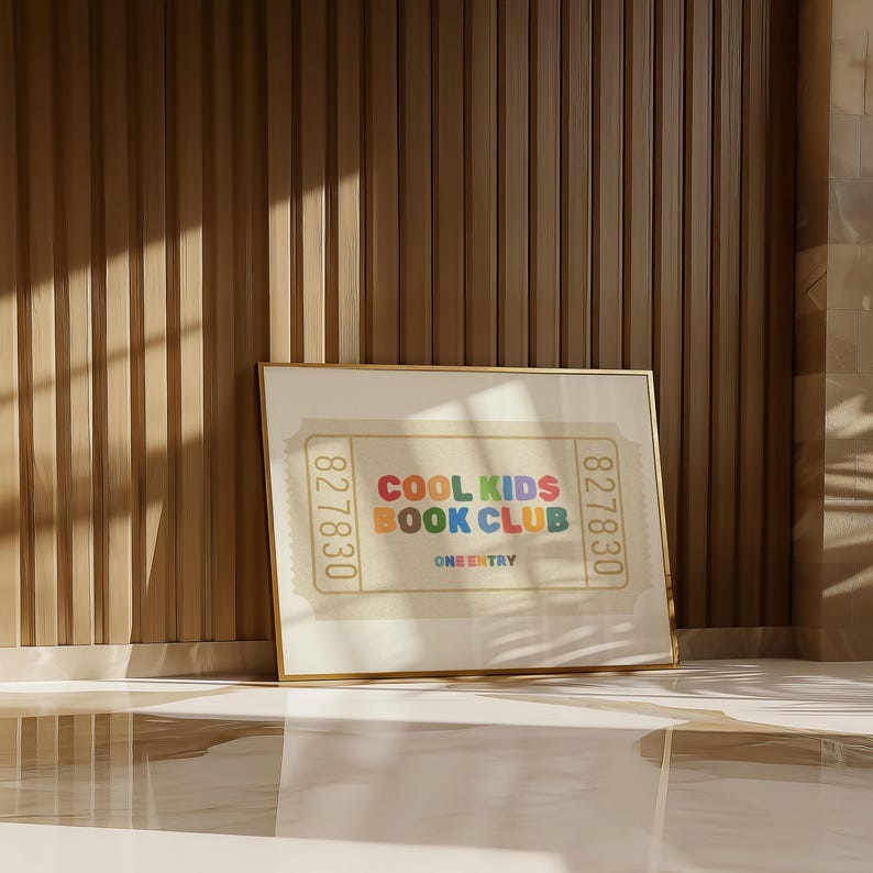 May include: A framed print with the text "COOL KIDS BOOK CLUB" in colorful letters. The print has a ticket design with the number "827830" on each side. The frame is gold, and the background is a light beige color.