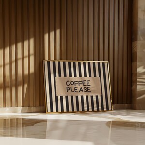Puede incluir: Una obra de arte enmarcada con un borde dorado presenta un fondo a rayas blancas y negras. Un parche rectangular beige en el centro muestra las palabras "COFFEE PLEASE" en may&uacute;sculas negras. La obra est&aacute; apoyada contra una pared de madera.