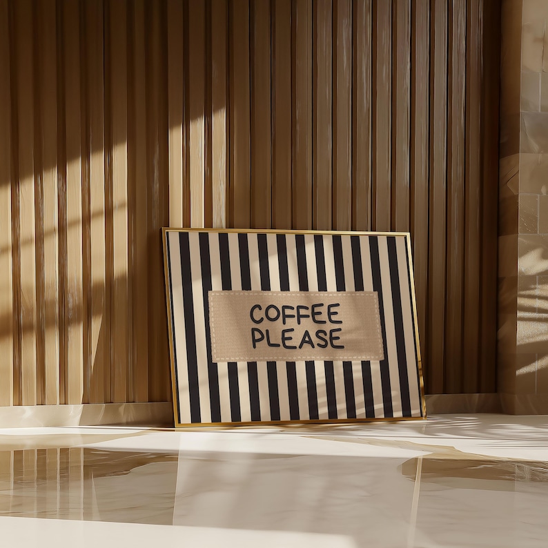 Puede incluir: Una obra de arte enmarcada con un borde dorado presenta un fondo a rayas blancas y negras. Un parche rectangular beige en el centro muestra las palabras "COFFEE PLEASE" en may&uacute;sculas negras. La obra est&aacute; apoyada contra una pared de madera.