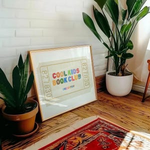 May include: A framed print with the text "COOL KIDS BOOK CLUB" in colorful letters. The print has a ticket design with the number "827830" on each side. The frame is gold, and the print is leaning against a white brick wall.