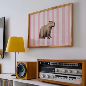 Op de afbeelding: Een ingelijste print van een capibara die op een roze en witte gestreepte achtergrond zit. De print hangt aan een witte muur boven een houten plank met een lamp, een luidspreker en een stereo.