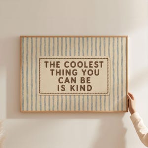 May include: A rectangular wall art print featuring a light beige background with vertical blue stripes. In the center, a rectangular patch with a stitched border displays the dark brown text: THE COOLEST THING YOU CAN BE IS KIND.