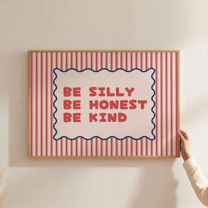 Op de afbeelding: Een ingelijste print met een rood-witte gestreepte achtergrond. De print heeft een witte, geschulpte vorm met de woorden "BE SILLY BE HONEST BE KIND" in rode tekst. De lijst is lichtbruin.