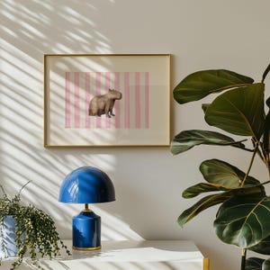 Op de afbeelding: Een ingelijste print van een capibara die op een roze-witte gestreepte achtergrond staat. Een blauwe lamp met een paddenstoelvormige kap staat op een wit oppervlak voor de print. Een groene, bladerrijke plant bevindt zich op de voorgrond.