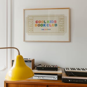May include: A framed print featuring a ticket design with the text "COOL KIDS BOOK CLUB" in colorful letters. The print is displayed above a wooden cabinet with a yellow lamp, a small amplifier, and electronic music equipment.