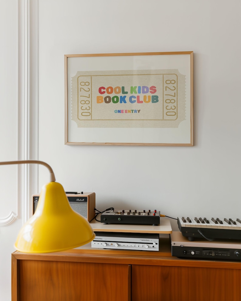 May include: A framed print featuring a ticket design with the text "COOL KIDS BOOK CLUB" in colorful letters. The print is displayed above a wooden cabinet with a yellow lamp, a small amplifier, and electronic music equipment.