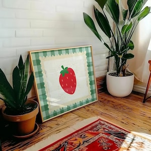 Puede incluir: Una impresi&oacute;n enmarcada con una fresa roja con un tallo verde. La obra de arte tiene un fondo blanco, un borde de cuadros verdes y un marco dorado. La impresi&oacute;n est&aacute; apoyada contra una pared de ladrillos blancos.