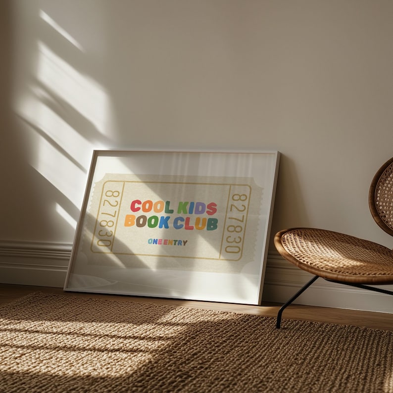 May include: Framed print featuring a ticket design with the text "COOL KIDS BOOK CLUB" in colorful letters. The print is displayed on a wall, next to a woven chair, and a brown rug.
