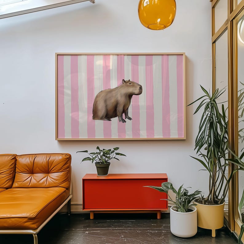 Op de afbeelding: Een ingelijste print van een capibara die op een roze en witte gestreepte achtergrond staat. De print hangt aan een witte muur boven een rode kast.