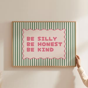 Puede incluir: Una impresión artística enmarcada con un fondo a rayas verdes y blancas. La impresión presenta un borde festoneado rosa con las palabras "BE SILLY BE HONEST BE KIND" en letras rosas. El marco es de color marrón claro.