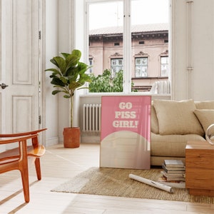 May include: A pink and white poster with the text "GO PISS GIRL!" in bold white letters. The poster is hanging on a white wall in a living room setting.