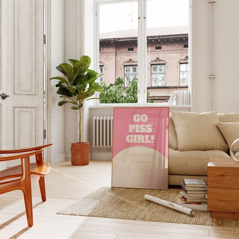 May include: A pink and white poster with the text "GO PISS GIRL!" in bold white letters. The poster is hanging on a white wall in a living room setting.
