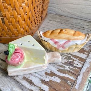 May include: Two ceramic salt and pepper shakers shaped like a slice of pie and a loaf of bread in a basket. The pie shaker has a red and green apple on top. The bread shaker has a red and white checkered pattern on the basket.