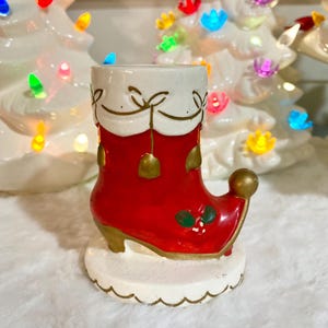 May include: A ceramic Christmas boot candle holder. The boot is red with gold accents, a white cuff, and a scalloped base. The boot has a gold heel and a small holly design. Festive holiday decor.