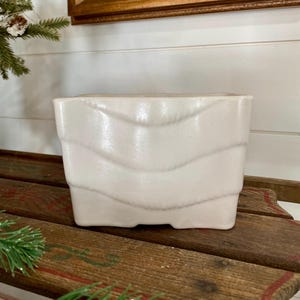 May include: White ceramic planter with a wavy design. The planter has a matte finish and is sitting on a wooden surface.