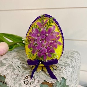 May include: A yellow Easter egg with purple and gold trim is decorated with purple flowers and gold beads. The egg is sitting on a stand with a purple ribbon.