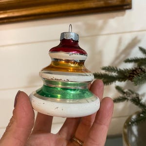 May include: A vintage Christmas tree ornament with a layered design. The ornament features a red top, gold center, and green base, with white and silver accents. The ornament is held in a hand.