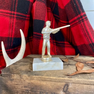 May include: A silver-toned figurine of a hunter wearing a cap and vest, holding a rifle in a shooting stance. The figure is mounted on a rectangular, off-white marble base with a smooth, polished finish, standing upright on a wooden surface.