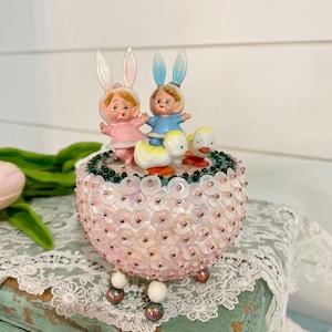 May include: A vintage Easter-themed trinket box with a pink and white beaded exterior. The box features three figurines: two children wearing bunny ears and one duck. The children are holding hands and the duck is sitting on the box.