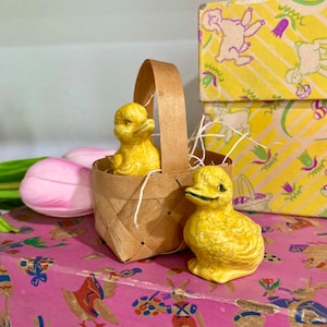 May include: A small woven basket with a handle holds two yellow ceramic ducklings, one inside and one outside. A third duckling stands beside the basket. The basket sits on a pink box with a colorful Easter-themed pattern. Yellow boxes with a similar pattern are in the background.